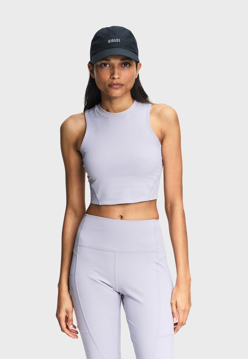 Femme portant une casquette noire et un haut court sans manches gris clair, avec un legging taille haute assorti, sur un fond uni.