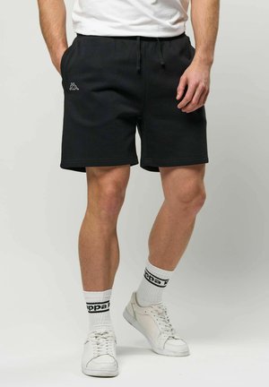 Bărbat purtând pantaloni scurți negri cu logo-ul Kappa, adidași albi și șosete albe până la gleznă cu branding negru Kappa, stând pe un fundal simplu.