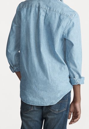 Persona que lleva una camisa azul claro de manga larga con puños remangados y vaqueros de mezclilla azul oscuro, vista desde atrás.