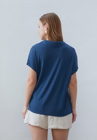 Navy modrý polo štýlový top s krátkymi rukávmi, jemnou textúrou a bočnými rozparkami. Skombinovaný s svetlými textúrovanými šortkami.