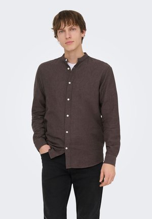 Ung mand med lys hud og brunt hår iført en mørkebrun skjorte med knapper, en hvid undertrøje og sorte bukser, stående med den ene hånd i lommen.