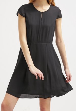 Kobieta w krótkiej, czarnej sukience do kolan z krótkimi rękawami, okrągłym dekoltem i małym wycięciem w kształcie klucza na wysokości piersi.