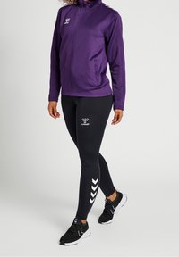Lila zip-up träningsjacka med hög krage, i kombination med svarta leggings som har en vit logotipo och ett chevronmönster på sidan. Svarta träningssneakers.
