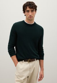 Maglione a girocollo lavorato a maglia verde scuro, abbinato a pantaloni beige e una cintura marrone con fibbia circolare. Tessuto morbido con polsini a coste.
