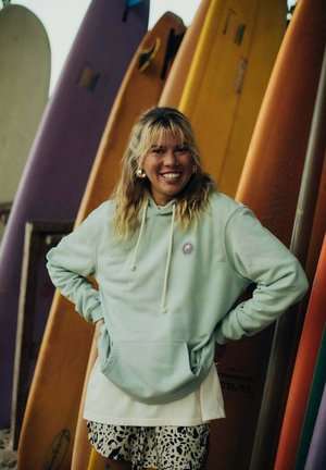 Miętowa bluza z kapturem z graficznym logo, nałożona na białą koszulę, zestawiona z czarno-białą spódnicą w wzory. W tle kolorowe deski surfingowe.