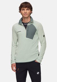Light green half-zip pullover with a gray chest panel, featuring a front zip pocket and logo on the sleeve, made from textured fabric.