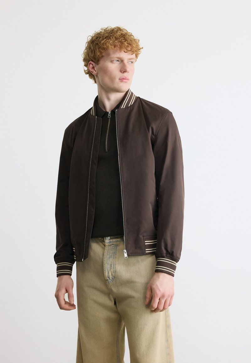 Νεαρός άνδρας με σγουρά μαλλιά, ντυμένος με σκούρο καφέ μπουφάν τύπου bomber με φερμουάρ, μαύρο πουκάμισο και μπεζ παντελόνι, κοιτάζοντας προς τα πλάγια μπροστά σε απλό φόντο.