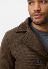 Brown textured coat with a notched collar, featuring a single black button and layered over a gray sweater. Close-up view of the neckline.