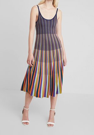 Femme portant une robe midi sans manches à rayures verticales multicolores avec des talons blancs à bride autour de la cheville, debout devant un fond uni.