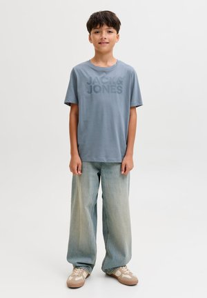 Jeune garçon debout, portant un t-shirt bleu "JACK & JONES", un jean large clair et des baskets beige sur un fond blanc uni.