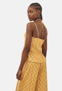 Top peplum amarillo con estampado, tirantes delgados y cintura fruncida, que presenta un diseño floral en verde y blanco sobre una tela texturizada.