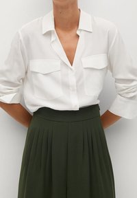 Chemise blanche à boutons avec deux poches poitrine, manches retroussées. Associée à un pantalon olive vert à taille haute et à pinces. Texture de tissu lisse.