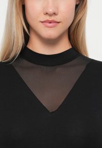 Black top featuring a high neckline and a sheer mesh inset in a V-shape at the chest, highlighting a smooth cotton blend material.