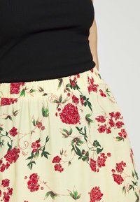 Květinová sukně s žlutým pozadím, zdobená červenými květy a zelenými listy. Elastický pas, lehký materiál a splývavá textura.