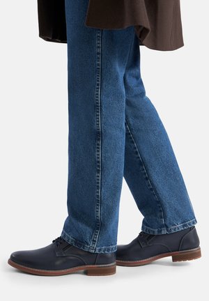 Tamsiai mėlyni odiniai batai su raišteliais ir lygiu paviršiumi, dėvimi su mėlynais džinsais. Turi rudą gumos pado dalį ir subtilias siuvimo detales.