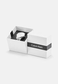Sølv rustfritt stål klokke med hvit skjerm, vist i en hvit eske med en svart rem og etikett med "Calvin Klein."