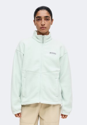 Person iført en lys mintgrøn Columbia fleecejakke med fuld lynlås og beige bukser, der står front mod en ensfarvet baggrund.