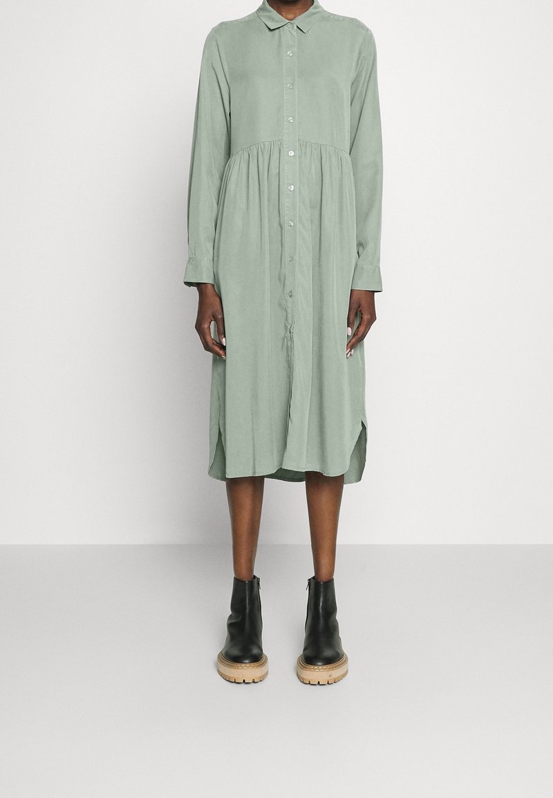 Sukienka z długim rękawem, z zapięciem na guziki, uszyta z miękkiego zielonego materiału, z marszczoną talią i bocznymi rozcięciami, w połączeniu z czarnymi botkami na lekki tle.