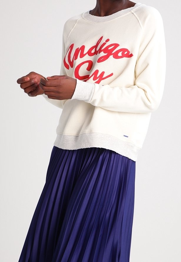 Sweatshirt crème arborant un texte rouge vif "Indigo", associé à une jupe plissée bleu marine. La tenue combine différentes textures et comprend un col côtelé.