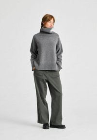 Maglione grigio oversize con collo a dolcevita e dettagli a costine, abbinato a pantaloni verdi a gamba larga. Scarpe nere completano l'outfit.