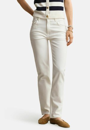 Frau trägt weiße Jeans mit geradem Bein, ein gestreiftes ärmelloses Oberteil, braune Slipper und Armbänder am rechten Handgelenk.