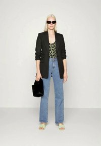 Mulher com cabelo loiro longo, vestindo um blazer preto, uma blusa de estampado de leopardo, jeans azuis, sandálias verdes, óculos de sol pretos, segurando uma bolsa preta.