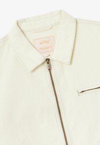 Veste beige clair en coton avec une texture lisse. Dotée d'une fermeture éclair, d'un col pointu et d'une poche poitrine zippée.