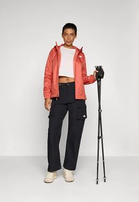 Lichtgewicht koraalkleurig windjack, wit crop topje, zwarte cargobroek met zakken, met wandelstokken in de hand, neutrale achtergrond.