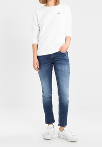 Sudadera blanca de cuello redondo con puños y dobladillo acanalados, combinada con jeans de denim azul. Las zapatillas blancas completan el atuendo. Detalle sutil del logo en la sudadera.