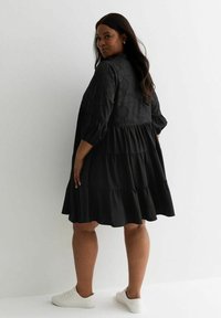 Vestido negro de corte en capas con mangas sueltas, que presenta detalles bordados en el corsé. El material parece suave; combinado con zapatillas blancas.