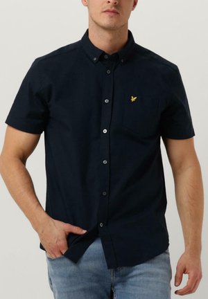 Mand iført en mørk marineblå kortærmet skjorte med knapper og et lille gult fuglelogo på brystet, kombineret med lyseblå jeans.