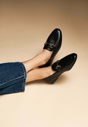 Mocassins en cuir noir avec un talon bas, présentant un détail décoratif en métal doré. Les chaussures ont une finition lisse et un design à bout pointu.