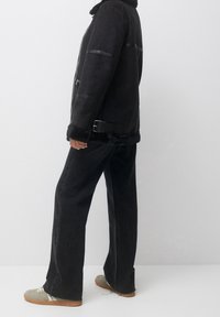 Veste en shearling noire avec des accents en cuir lisse, dotée d'une taille à boucle, portée avec un jean large noir et des baskets grises.