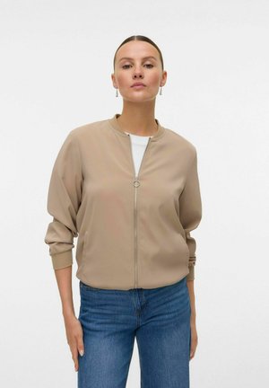 Beige bomberjack met een rits aan de voorkant, geribde manchetten en een ronde halslijn, gedragen met blauwe jeans en een wit shirt eronder.