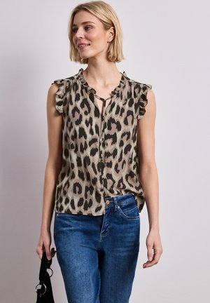 Blonde Frau trägt ärmellose Bluse mit Leopardenmuster und blaue Jeans, hält eine schwarze Handtasche vor einem schlichten hellen Hintergrund.