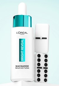 Sérum Bright Reveal Niacinamide de L'Oréal Paris dans un flacon en verre blanc avec un compte-gouttes turquoise, accompagné d'objets de couleur blanche et noire.