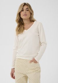 Light beige long-sleeve V-neck top with a scalloped edge. Soft texture, paired with cream-colored pants featuring black polka dots.