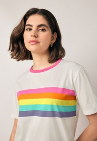 Rövid ujjú póló világos bézs színben, élénk vízszintes csíkokkal rózsaszín, narancssárga, sárga, zöld, türkiz és lila színekben. Kerek nyakkivágás.