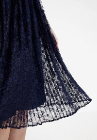 Granatowa koronkowa sukienka z plisowanym wzorem; przezroczysty materiał z kwiatowymi deserami; warstwowy dół; zwiewny krój; ręka delikatnie trzymająca materiał.