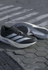 Paire de chaussures de course Adidas Adizero noir et blanc placée sur un trottoir gris texturé près d’un passage piéton.