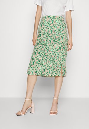Saia midi floral com fundo verde, apresentando pequenas flores cor-de-rosa e brancas, fenda lateral e cintura ajustada. Usada com saltos bloco cor-de-rosa.