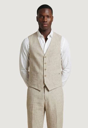 Uomo che indossa un gilet beige in lino e pantaloni abbinati con una camicia bianca su uno sfondo semplice.