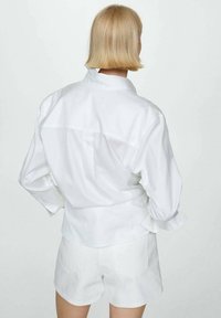 Camisa blanca de botones con mangas largas y un detalle de pliegue en la parte posterior, combinada con pantalones cortos blancos. La tela parece suave y ligera.
