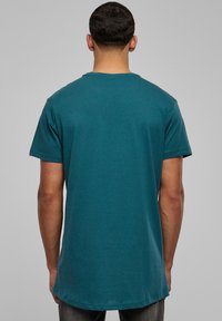 Tricou scurt, de culoare teal, cu mâneci scurte și decolteu rotund. Vedere din spate arată un croi drept și o margine ușor mai lungă. Fabricat dintr-un material moale cu o textură netedă.