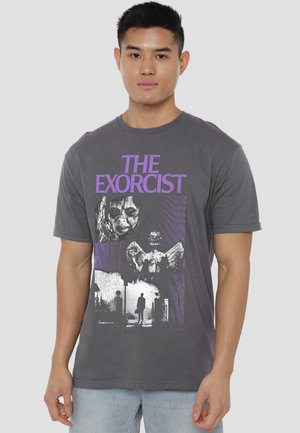 Young man wearing dark gray T-shirt featuring purple "The Exorcist" text, a scary face, angel statue, and shadowy figure in fog.