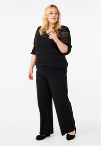 Zwart kanten top met patroon, driekwart mouwen, gecombineerd met zwarte wijde broek en zwarte ballerina's met strikken. Witte achtergrond.