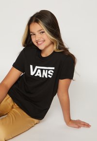 Camiseta de algodón negra con logo blanco de "VANS". El modelo está sentado en el suelo, vistiendo pantalones dorados. Textura suave y corte relajado.