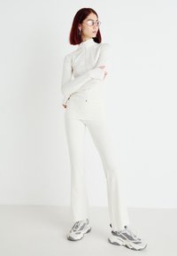 White fitted tracksuit with a high collar and zip closure, long sleeves, and flared pants, paired with grey chunky sneakers.