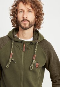 Olive green hoodie with gray sleeves, textured fabric, adjustable drawstrings, zip closure, and a small logo patch on the front.