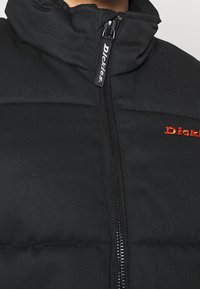 Doudoune noire avec col montant, dotée d'une fermeture éclair avant et d'une broderie rouge du logo « Dickies » sur la poitrine. Tissu à texture.